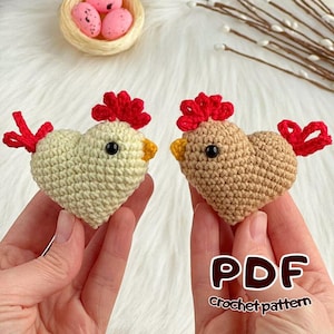 Peut inclure: Deux poulets en crochet en forme de cœur, crème et beige, avec des crêtes et des plumes de queue rouges et des becs jaunes. Les poulets ont des yeux noirs. L'image montre également un nid avec des œufs roses tachetés et le texte "PDF Crochet pattern".