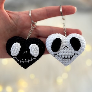 May include: Two crocheted heart-shaped keychains with skull faces. One keychain is black with white eyes and the other is white with black eyes.