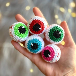 May include: Five crocheted eyeball ornaments in different colors, including red, green, blue, and pink. Each eyeball has a black pupil and white sclera.
