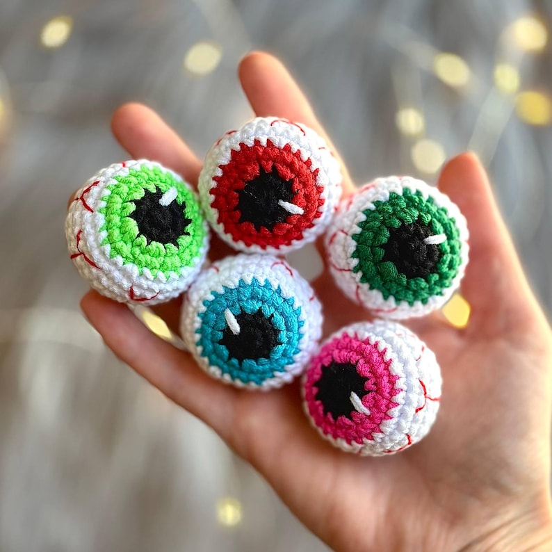 May include: Five crocheted eyeball ornaments in different colors, including red, green, blue, and pink. Each eyeball has a black pupil and white sclera.