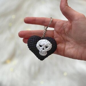 Puede incluir: Un llavero negro en forma de coraz&oacute;n tejido a ganchillo con un dise&ntilde;o de calavera blanca. El llavero hecho a mano est&aacute; sostenido por una mano, mostrando los intrincados detalles. La calavera est&aacute; centrada en el coraz&oacute;n, creando una est&eacute;tica g&oacute;tica.