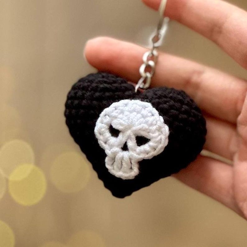 Puede incluir: Un llavero en forma de coraz&oacute;n negro tejido a ganchillo con un dise&ntilde;o de calavera blanca. La calavera est&aacute; centrada en el coraz&oacute;n y el llavero tiene una cadena de metal plateado. El fondo est&aacute; borroso.