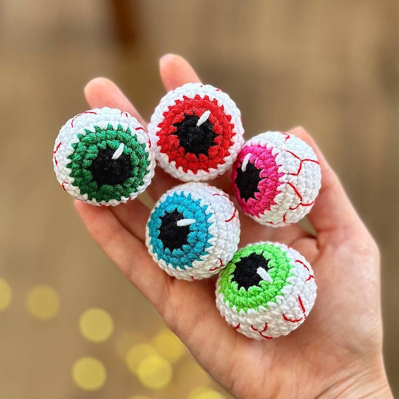 May include: Five crocheted eyeball ornaments in different colors: green, red, pink, blue, and green. The eyeballs are white with black pupils and red veins.