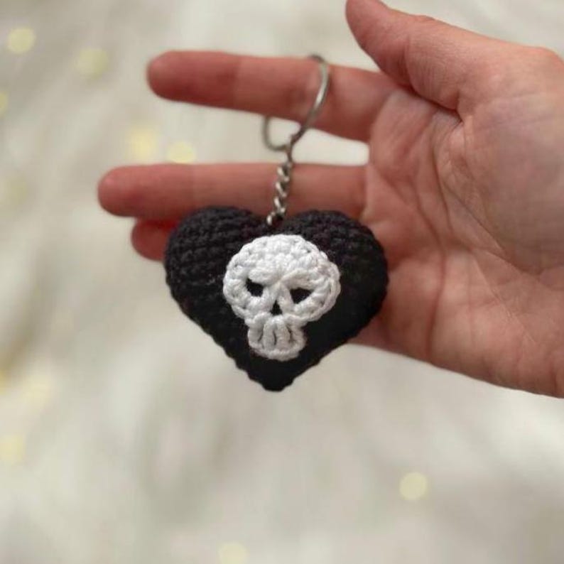 Puede incluir: Un llavero en forma de coraz&oacute;n negro tejido a ganchillo con un dise&ntilde;o de calavera blanca. El llavero est&aacute; hecho de hilo y tiene un anillo de metal plateado. La calavera est&aacute; centrada en el coraz&oacute;n.