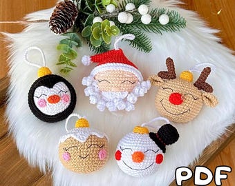 May include: Five crocheted Christmas ornaments, including a penguin, Santa Claus, reindeer, gingerbread person, and snowman. Each ornament has a smiling face and is decorated with details like a red nose, a white beard, and a black top hat. The ornaments are displayed on a white fluffy surface with a green sprig of greenery.