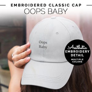 Può includere: Cappellino da baseball ricamato di colore grigio con il testo "Oops Baby" in ricamo nero. Il cappellino ha un cinturino regolabile sul retro.