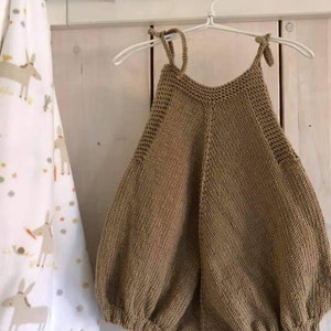 May include: A brown knitted baby onesie with straps hanging on a white hanger. The onesie has a loose fit and is made of a soft, textured fabric.