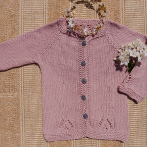 Może przedstawiać: Różowy kardigan dzianinowy z zapięciem na guziki i delikatnym wzorem koronkowym na dolnym brzegu. Kardigan leży płasko na tkanej powierzchni.