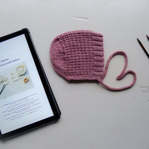 Puede incluir: Un gorro de bebé de punto rosa con un lazo, agujas de tejer, tijeras y una tableta digital que muestra un patrón de tejido. El patrón se titula "Anleitung: Babymat  when for quilts  baby jumpers".