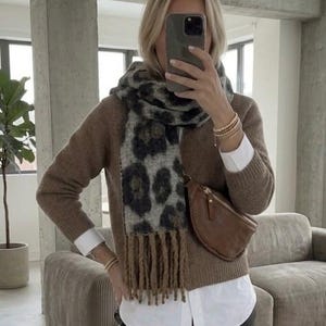 Könnte beinhalten: Eine braune Strickjacke über einem weißen Hemd mit Kragen und einer schwarzen Hose. Ein grauer und schwarzer Schal mit Leopardenmuster und Fransen liegt um den Hals. Eine braune Umhängetasche aus Leder wird quer über der Brust getragen.