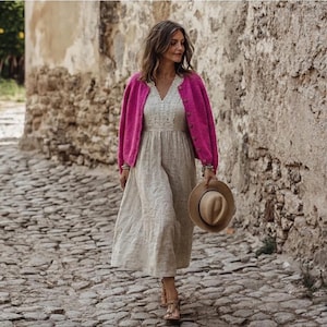 Könnte beinhalten: Ein beiges Midikleid mit weißem Spitzenoberteil und V-Ausschnitt, kombiniert mit einer leuchtend pinkfarbenen, langärmeligen Strickjacke mit Knöpfen. Das Outfit wird durch einen beigen Hut mit breiter Krempe und braune Riemchensandalen ergänzt.