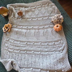 Könnte beinhalten: Eine hellgraue Strickdecke mit verschiedenen strukturierten Mustern, darunter Noppen, Zopfmuster und Herzformen. Die Decke ist mit kleinen Pilzfiguren aus Holz, einem Holzherz, einem Plüschigel und einem kleinen orangefarbenen Kürbis verziert.