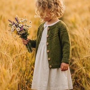 Könnte beinhalten: Eine langärmelige, olivgrüne Strickjacke mit Rundhalsausschnitt und Holzknöpfen, getragen über einem schlichten cremefarbenen Kleid. Die Strickjacke hat ein strukturiertes Strickmuster an der Passe und gerippte Bündchen, kombiniert mit braunen Lederschuhen.