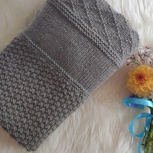 Könnte beinhalten: Eine gefaltete, graue Strickdecke mit drei deutlich strukturierten Abschnitten. Ein kleines braunes Lederlesezeichen liegt in der Nähe der Decke. Eine Vase mit Blumen mit gelben, rosa und weißen Blüten ist mit einem blauen Band gebunden.