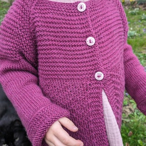 Puede incluir: Un cárdigan de punto rosa con cierre de botones. El cárdigan tiene un patrón de punto texturizado y tres botones con diseños florales.