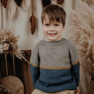Puede incluir: Un niño pequeño con un suéter a rayas gris, azul y marrón se sienta en un taburete de madera. Está sonriendo y mirando a la cámara. El fondo es una pared marrón claro con una variedad de artículos decorativos.