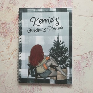 May include: A black and white checked planner with the text "Kerrie's Christmas Planner" on the cover. The cover also features a cartoon illustration of a woman sitting in a chair with a Christmas tree behind her. The text "Merry Christmas" is written on the tree.
