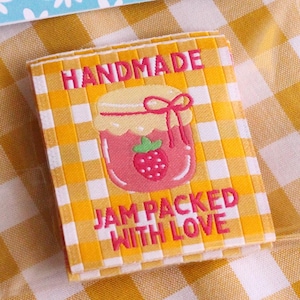 Könnte beinhalten: Näh-Etiketten mit dem Text "THE COSY CLUB HANDMADE" und "BRIGHT & CHEERFUL SEWING LABELS" auf blauem floralem Hintergrund. Ein zweites Etikett lautet "HANDMADE JAM PACKED WITH LOVE" mit einer Marmeladenglas-Grafik. Die Etiketten befinden sich auf einem gelb-weiß karierten Hintergrund.