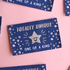 May include: Navy blue rectangular labels with the text "TOTALLY UNIQUE" and "ONE OF A KIND" in silver. Each label features a smiling star and star accents. The labels are scattered on a pink background.