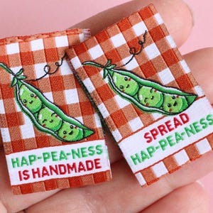 May include: Two small fabric labels with a red and white checkered pattern. Each label features an embroidered green pea pod with smiling peas. One reads "HAP-PEA-NESS IS HANDMADE," the other says "SPREAD HAP-PEA-NESS."