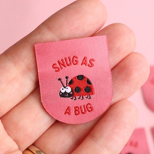 Peut inclure: Étiquette en tissu rose avec le texte "SNUG AS A BUG" et une illustration de coccinelle de dessin animé. La coccinelle est rouge avec des points noirs et une tête noire. Plusieurs étiquettes similaires sont visibles.