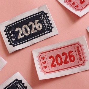 May include: A collection of fabric labels, each embroidered with the year "2026". Some labels feature a navy blue border with silver text, while others are red with a heart design. The word "HANDMADE" is also displayed on the labels.