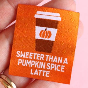 May include: Orange fabric label with a coffee cup graphic and a pumpkin. The text reads "Sweeter Than a Pumpkin Spice Latte". The label is held in a hand against a pink background.