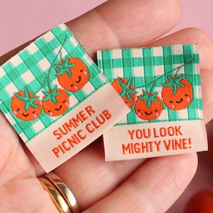 Könnte beinhalten: Zwei kleine Stoffetiketten mit Tomatenillustrationen und Text. Auf einem steht "Summer Picnic Club" und auf dem anderen "You Look Mighty Vine!" Die Etiketten werden in einer Hand gehalten, wobei ein goldener Ring sichtbar ist. Im Hintergrund befindet sich ein Strauch mit roten Tomaten.