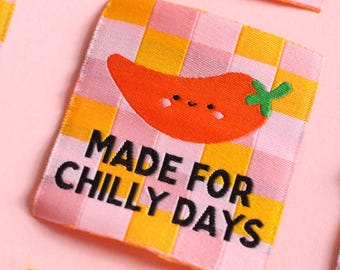 Könnte beinhalten: Nahaufnahme mehrerer Stoffetiketten mit einem rosa, gelben und weißen Karomuster. Jedes Etikett zeigt eine orangefarbene Chili-Pfeffer-Illustration und den Text "MADE FOR CHILLY DAYS" in Schwarz.