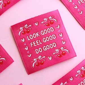 Op de afbeelding: Roze stoffen labels met de tekst "LOOK GOOD FEEL GOOD DO GOOD" in het wit. De labels bevatten illustraties van kersen en strikken. De labels zijn verspreid op een roze achtergrond.
