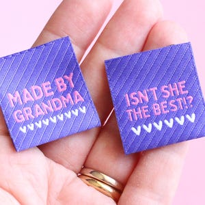 May include: Two square purple fabric labels with diagonal lines and pink text. One says "MADE BY GRANDMA" and the other says "ISN'T SHE THE BEST?". Both have a white zigzag design at the bottom. The labels are held in a hand.