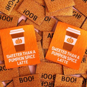 May include: Two rectangular orange tags featuring a brown coffee cup graphic with a white pumpkin icon and the text SWEETER THAN A PUMPKIN SPICE LATTE, resting on a pile of glittery copper-colored tags printed with the words BOO and ARGH.