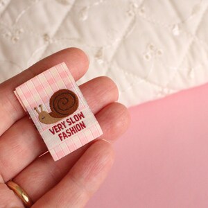 Peut inclure: Une &eacute;tiquette en tissu vichy rose et blanc avec une illustration de limace brune et le texte "Very Slow Fashion".