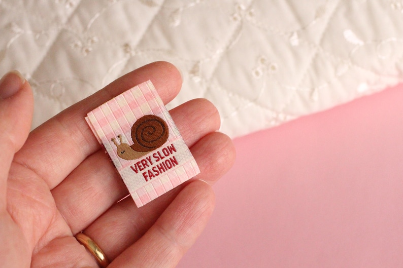 Peut inclure: Une &eacute;tiquette en tissu vichy rose et blanc avec une illustration de limace brune et le texte "Very Slow Fashion".