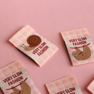 Peut inclure: Cinq &eacute;tiquettes en tissu vichy rose et blanc avec le texte "Very Slow Fashion" et une image brod&eacute;e d'un escargot ou d'un paresseux.