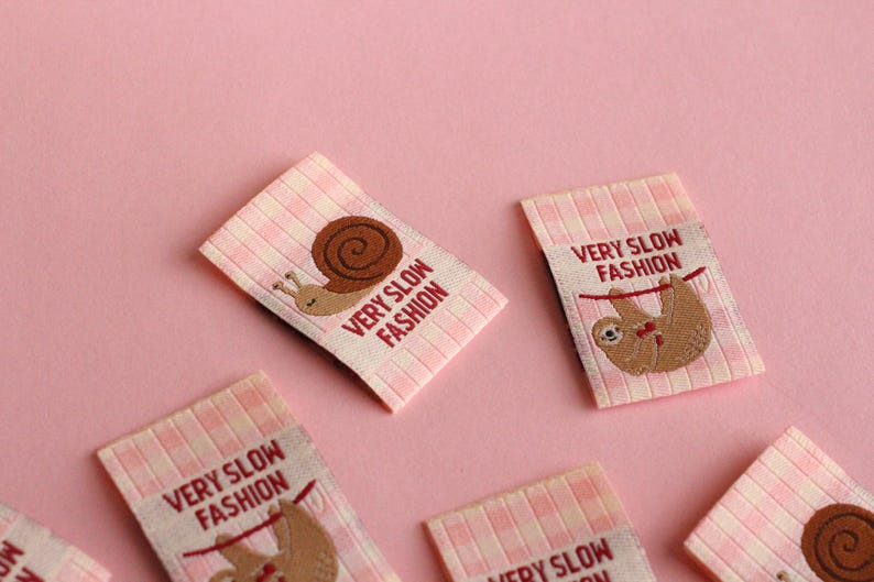 Peut inclure: Cinq &eacute;tiquettes en tissu vichy rose et blanc avec le texte "Very Slow Fashion" et une image brod&eacute;e d'un escargot ou d'un paresseux.