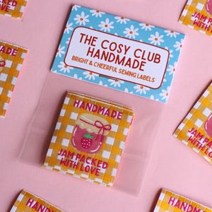 May include: A collection of sewing labels with a gingham pattern and a strawberry jam jar illustration. The labels read "Handmade" and "Jam Packed With Love." A blue and white floral card reads "The Cosy Club Handmade."
