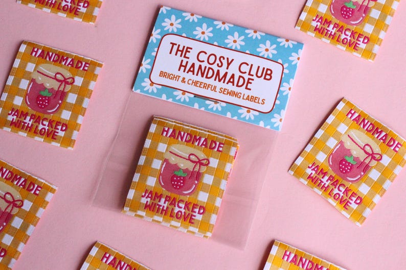 May include: A collection of sewing labels with a gingham pattern and a strawberry jam jar illustration. The labels read "Handmade" and "Jam Packed With Love." A blue and white floral card reads "The Cosy Club Handmade."