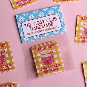 May include: A collection of handmade sewing labels with a gingham pattern and a jar of jam illustration. The labels read "Handmade" and "Jam Packed With Love." A card with the text "The Cosy Club Handmade" is also visible.