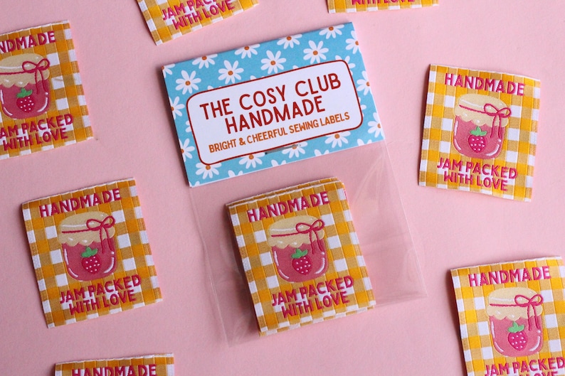 May include: A collection of handmade sewing labels with a gingham pattern and a jar of jam illustration. The labels read "Handmade" and "Jam Packed With Love." A card with the text "The Cosy Club Handmade" is also visible.