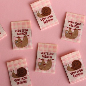 Peut inclure: Huit &eacute;tiquettes en tissu vichy rose et blanc avec le texte "VERY SLOW FASHION" et une image brod&eacute;e d'un paresseux ou d'un escargot.