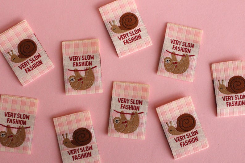 Peut inclure: Huit &eacute;tiquettes en tissu vichy rose et blanc avec le texte "VERY SLOW FASHION" et une image brod&eacute;e d'un paresseux ou d'un escargot.