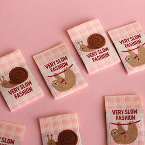Peut inclure: Six &eacute;tiquettes en tissu vichy rose et blanc avec le texte "VERY SLOW FASHION" et une image brod&eacute;e d'un paresseux ou d'un escargot.