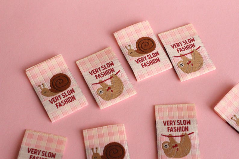Peut inclure: Six &eacute;tiquettes en tissu vichy rose et blanc avec le texte "VERY SLOW FASHION" et une image brod&eacute;e d'un paresseux ou d'un escargot.