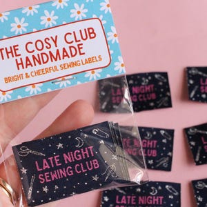 May include: A hand holds a packet of sewing labels against a pink backdrop. The top label reads "THE COSY CLUB HANDMADE" with a daisy pattern. The labels in the packet and scattered around say "LATE NIGHT SEWING CLUB" in pink on a navy background.