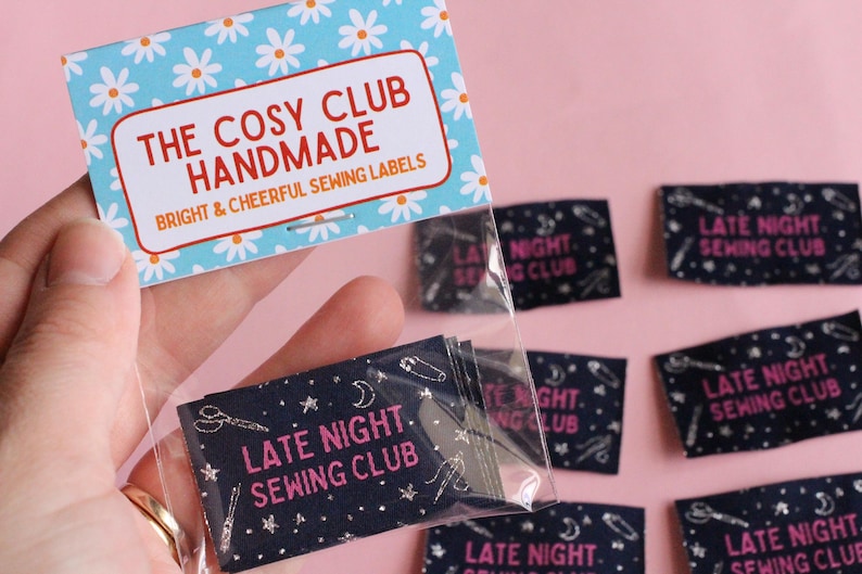 May include: A hand holds a packet of sewing labels against a pink backdrop. The top label reads "THE COSY CLUB HANDMADE" with a daisy pattern. The labels in the packet and scattered around say "LATE NIGHT SEWING CLUB" in pink on a navy background.