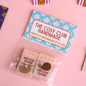 Peut inclure: Un ensemble de quatre &eacute;tiquettes en tissu thermocollantes &agrave; motif vichy rose et blanc. Les &eacute;tiquettes pr&eacute;sentent un paresseux, un escargot, une tasse de th&eacute; et un motif de marguerite. Les &eacute;tiquettes sont emball&eacute;es dans un sac en plastique transparent avec une carte bleue et blanche qui indique "The Cosy Club Handmade" et "thecosyclubhandmade.etsy.com".