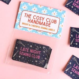 May include: Sewing labels with the text "LATE NIGHT SEWING CLUB" in pink, featuring illustrations of scissors, needles, and stars on a dark blue fabric. The labels are packaged with a card that reads "THE COSY CLUB HANDMADE".