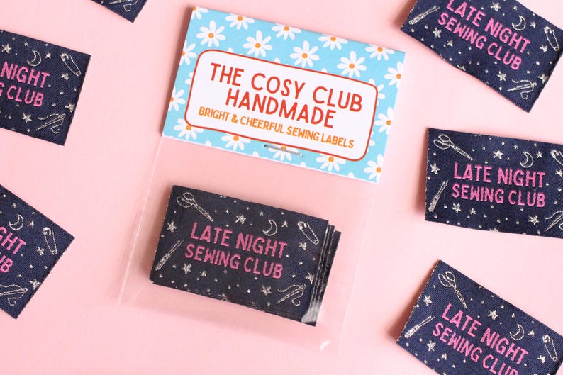May include: Sewing labels with the text "LATE NIGHT SEWING CLUB" in pink, featuring illustrations of scissors, needles, and stars on a dark blue fabric. The labels are packaged with a card that reads "THE COSY CLUB HANDMADE".