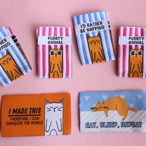May include: A collection of cat-themed fabric labels with various designs and text. Some labels read "I'D RATHER BE NAPPING" and "PURRTY ANIMAL". Others say "I MADE THIS THEREFORE, I CAN CONQUER THE WORLD" and "EAT, SLEEP, REPEAT".